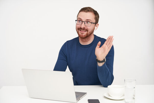 积极开朗的年轻英俊的金发男子微笑着高兴地举起手来打招呼 坐在白色背景的桌子旁 手里拿着笔记本电脑男性坐着姿势