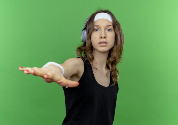 冷静身穿黑色运动服 头戴耳机 手举绿墙 做着冷静的手势的年轻健身女孩站立耳机运动装