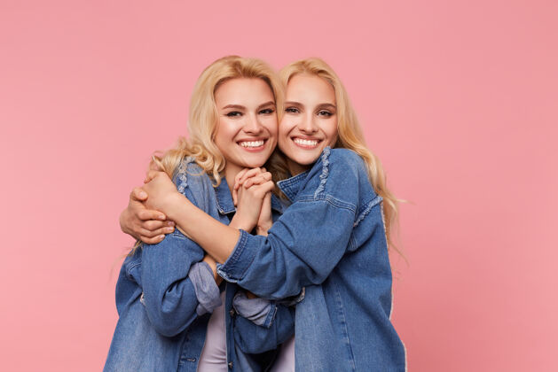 室内摄影棚的照片 年轻的快乐可爱的金发姐妹长波浪发看着相机迷人的微笑 同时拥抱可爱 站在粉红色的背景T恤白色欢呼