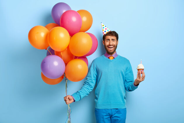 纸杯蛋糕半身镜头帅哥戴着生日帽和气球摆着蓝色毛衣男甜点休闲