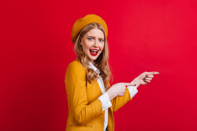 金发放松的法国女人用手指指着穿着考究的金发女孩站在红墙上情感服装优雅