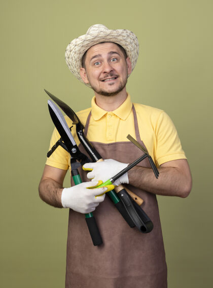 穿年轻的园丁 穿着连体衣 戴着帽子 戴着工作手套 手里拿着园艺设备 脸上带着微笑站连身衣工作