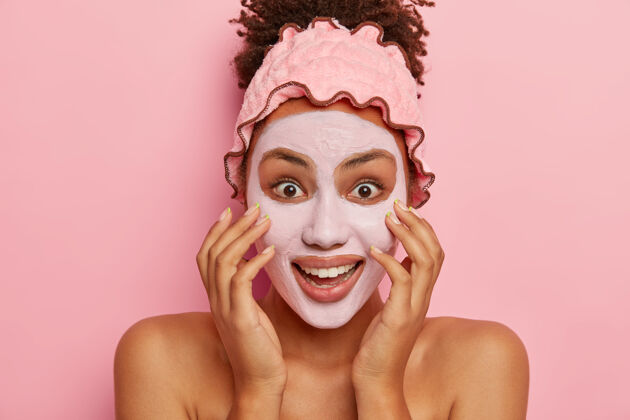 面膜快乐黑皮肤女人头像涂泥面膜 减少毛孔的能见度 享受美容产品深入肌肤的有效效果 站在室内粉色墙壁上露肩痘痘年轻快乐