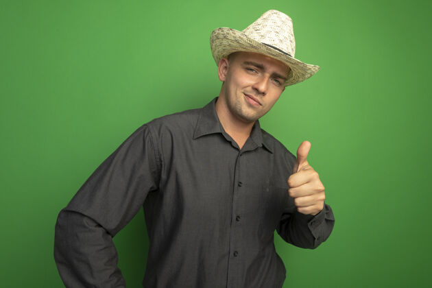 人身穿灰色衬衫 头戴夏帽的年轻帅哥站在绿色的墙上 面带笑容 竖起大拇指 望着前面人拇指姿势