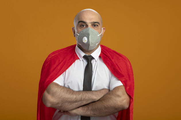 保护超级英雄的商人在保护面罩和红色披肩看着前面 双手交叉胸前 严重皱眉头站在橙色墙脸人工作