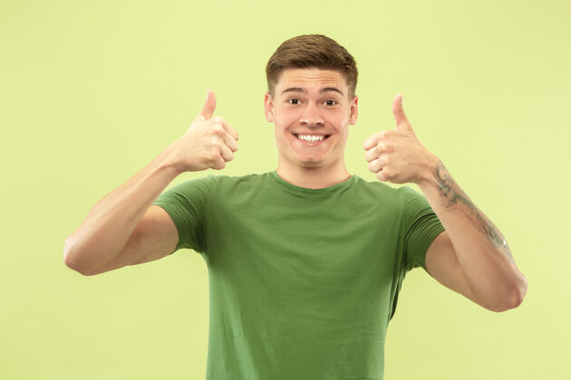 帅气绿色工作室背景上的白人年轻人半身肖像穿着衬衫的漂亮男模人类情感的概念 面部表情 销售 广告微笑 竖起大拇指成功手势衬衫