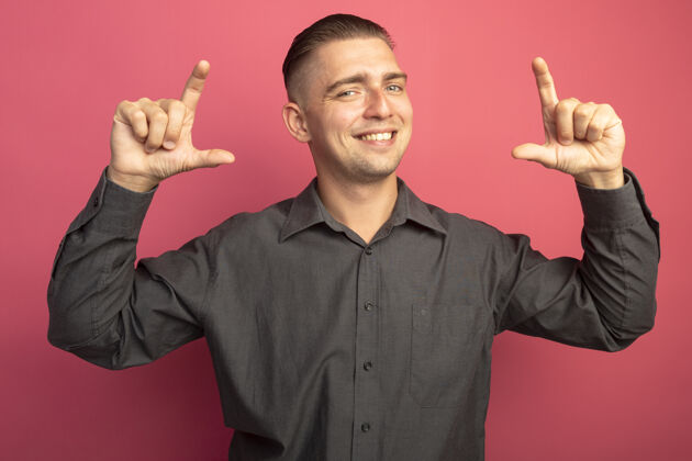 人身着灰色衬衫的年轻帅哥笑容可掬地展示着手指的尺码手势站在粉色的墙上人符号手指