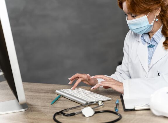 职业侧视图：女医生戴着医用面罩坐在办公桌旁 在键盘上写字面罩卫生工作者复印空间