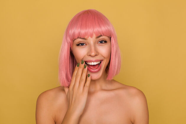 手室内照片 可爱 快乐 年轻的蓝眼睛女士 粉色的鲍勃发型 快乐地将手掌举到脸上 在芥末墙上摆着姿势 面带微笑女性姿势肖像