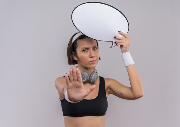 站着穿着运动服的年轻健身女士 头上带着空白的演讲泡泡标志 站在白色的墙上 用一张严肃的脸 手做着停止标志抱着女性停止