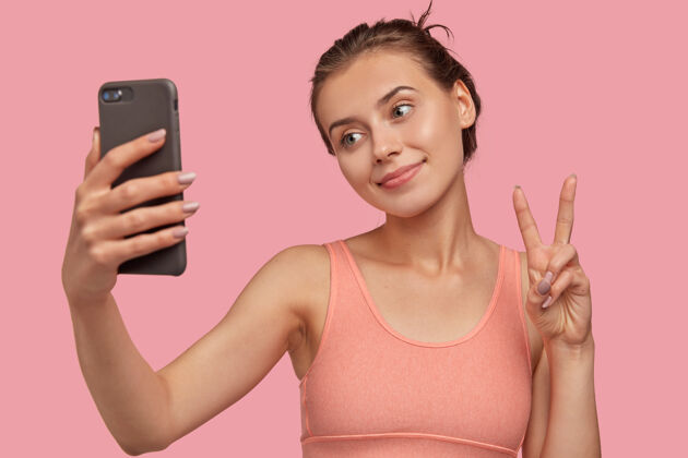 情绪积极的白人女性形象 皮肤清新 手持现代智能手机在前面 做和平的标志 歪着头 穿着休闲背心模特在粉红色的墙上漂亮女孩自拍肖像满意时尚手机
