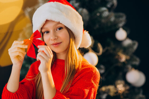 人可爱的少女戴着圣诞树旁的红色圣诞帽圣诞老人圣诞树红帽子