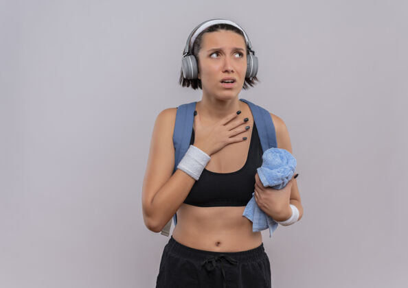 运动员年轻的健身女士穿着运动服 背着背包 头上戴着耳机 手放在胸前拿着毛巾 站在白色的墙上焦急地看着一边靠边站健康看