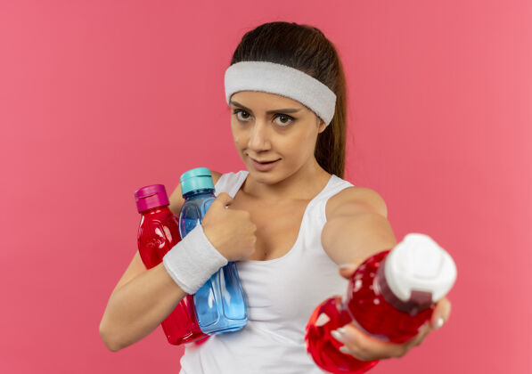 头带穿着运动服的年轻健身女 头上戴着一条带子 手里拿着几瓶水 站在粉红色的墙上 给其中一瓶水喝公动装持有
