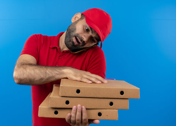 叠着一个留着胡须 身穿红色制服 戴着帽子的送货员站在蓝色的墙上 手里拿着一叠比萨饼盒 用手机聊天盒子帽子拿着
