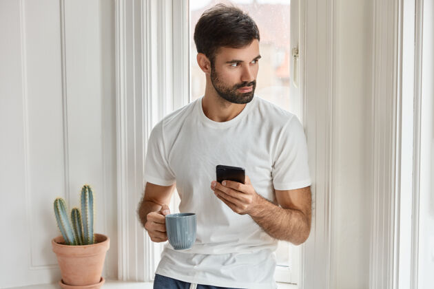 设备一个留着胡须的白人男子 穿着白色t恤 拿着手机和咖啡杯 安装新的应用程序 享受免费互联网 专注在一边 在餐厅点餐 喝咖啡胡须小玩意成人