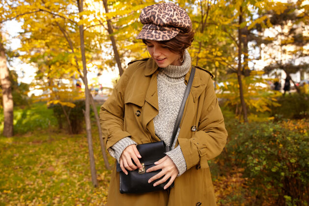 户外在温暖的秋日里 一位年轻时尚的黑发女士站在城市花园的上空 一边在她的黑色皮包里找东西风衣心情包