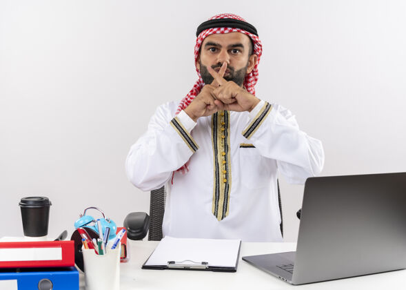 传统身着传统服装的阿拉伯商人坐在桌旁 用笔记本电脑交叉食指做着沉默的手势在办公室工作制造商务人士坐着