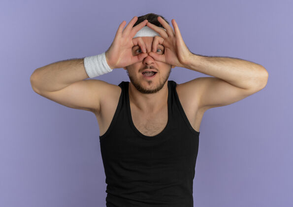 运动员戴着头巾的年轻健身男子做着双目手势 手指透过站在紫色墙上的手指望着前方人手势头带