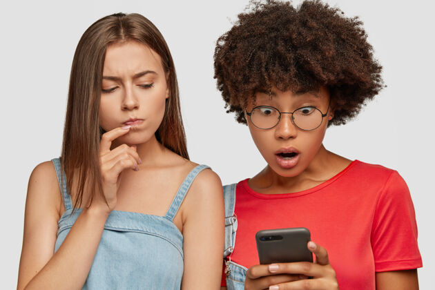 惊人照片中惊讶的非洲裔美国人和白人妇女看起来惊讶和怀疑进入智能手机一起手机困惑