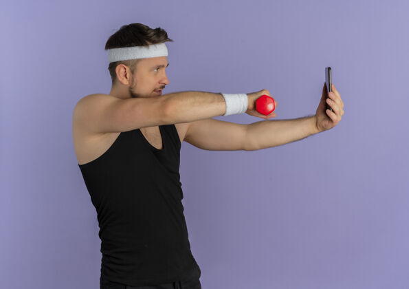 自拍戴着头巾的年轻健身男子用智能手机自拍 手里拿着哑铃站在紫色的墙上头带人人