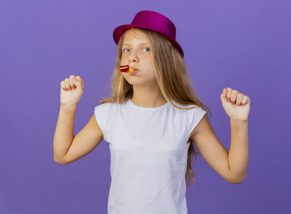 小美丽的小女孩在节日帽子吹哨子握紧拳头高兴和兴奋 生日派对的概念站在紫色的背景哨子紫色派对
