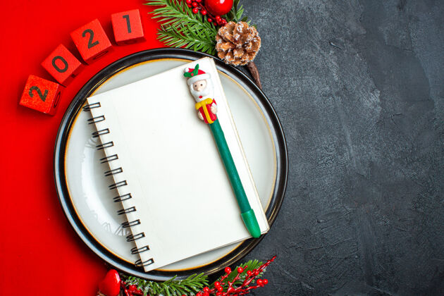 螺旋新年背景俯视图 餐盘上有螺旋笔记本 装饰配件杉木树枝 黑色桌子上的红色餐巾上有数字橄榄球球杉木餐盘