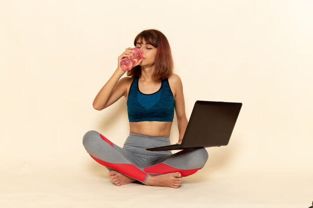 成人穿着蓝色衬衫的年轻女性的前视图 她使用笔记本电脑和白墙上的饮用水锻炼衬衫前面