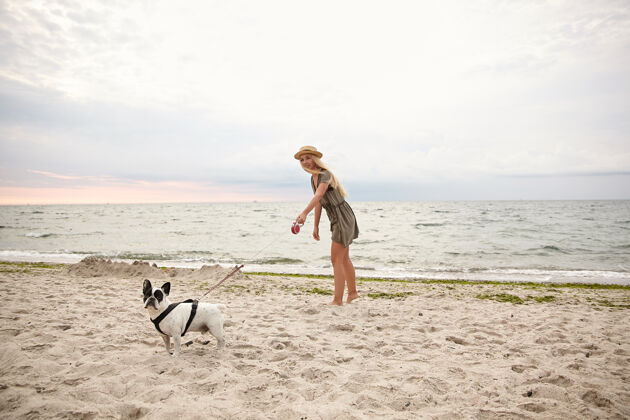 视图一个漂亮的金发年轻女子穿着夏装 戴着草帽 带着她的狗在阴天的沙滩上散步的水平户外镜头多云长白色