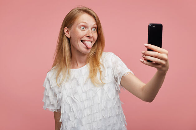 高兴有趣的照片 年轻快乐的红发女士伸出舌头和圆绿色灰色的眼睛 同时与她的手机 孤立在粉红色的背景照片自己雀斑欢呼手