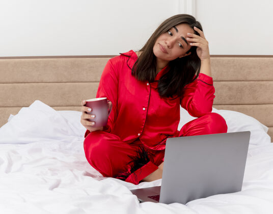 不高兴穿着红色睡衣的年轻漂亮女人坐在床上 手里拿着笔记本电脑和一杯咖啡 在浅色背景下的卧室里显得困惑和不安困惑睡衣咖啡