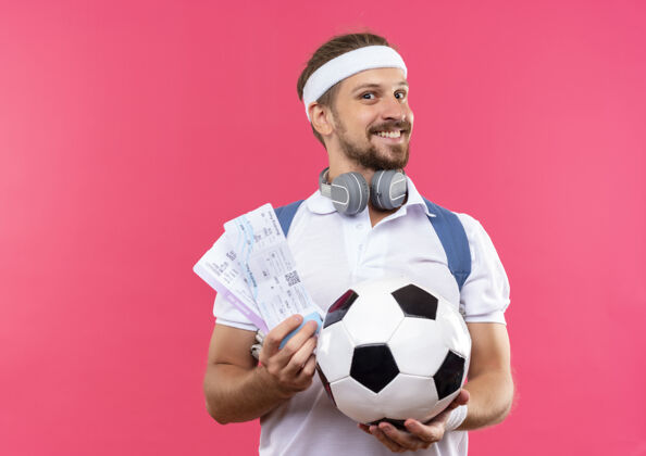 举行面带微笑的年轻帅气的运动型男人戴着头带和腕带 脖子上背着一个带耳机的包 手里拿着足球和机票 被隔离在粉红色的空间里粉色足球太空