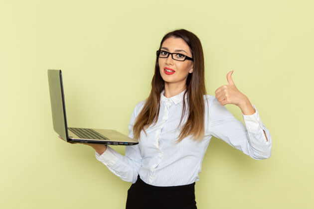 女士穿着白衬衫和黑裙子的女上班族在绿墙上拿着笔记本电脑的正视图漂亮工人拿着