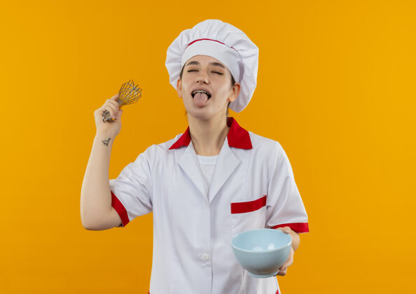 制服顽皮的年轻漂亮厨师 身着厨师制服 手里拿着打蛋器和碗 闭着眼睛闭着舌头 孤立地站在橙色的空间里漂亮橙色厨师