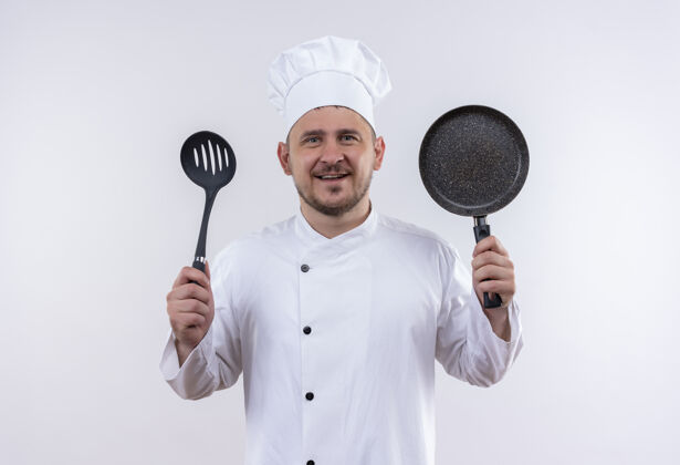 厨师面带微笑的年轻帅哥厨师身着厨师制服 拿着开槽汤匙和煎锅站在与世隔绝的白色空间里勺子厨师油炸