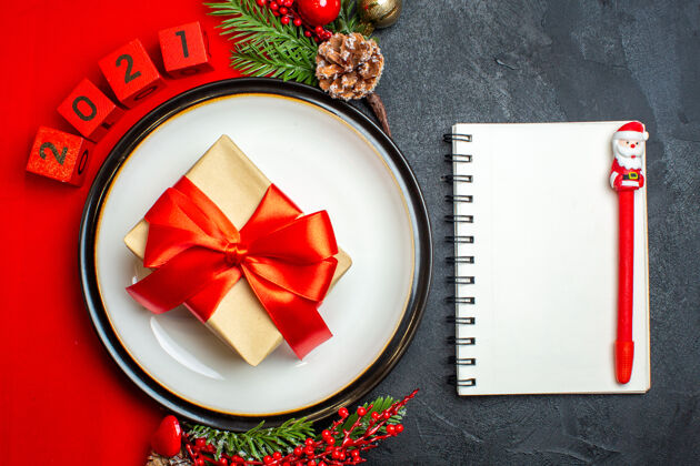 笔记本新年背景的顶视图 餐盘上有礼物装饰配件 红色餐巾上有杉木树枝和数字 黑色桌子上有笔的笔记本顶部餐巾钢笔