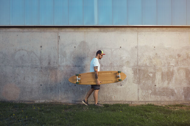 木头一个留着胡须和纹身的年轻人 穿着一件纯白的t恤 短裤 运动鞋和棒球帽 手里拿着一块长板 沿着水泥墙走专业滑板文化
