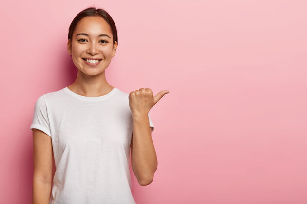指向快乐的亚洲年轻女子的水平镜头指向复制空间 展示一些好东西 穿白色t恤 帮助挑选最佳选择 推荐产品 粉色墙上的模特积极服装年轻
