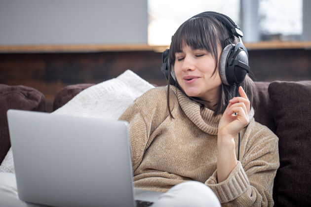 音乐美丽的女人戴着耳机在家里用笔记本电脑坐在沙发上听音乐听唱歌年轻