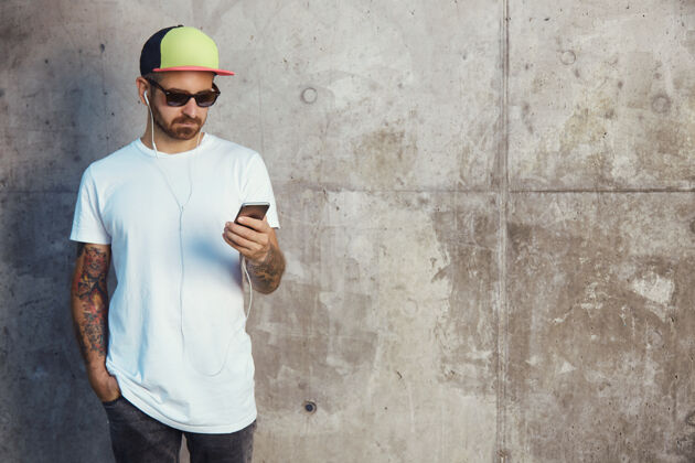 耳机一个戴着棒球帽 戴着太阳镜 穿着白色空白t恤的年轻人站在灰色水泥墙旁边 用智能手机读着什么应用程序视频休闲