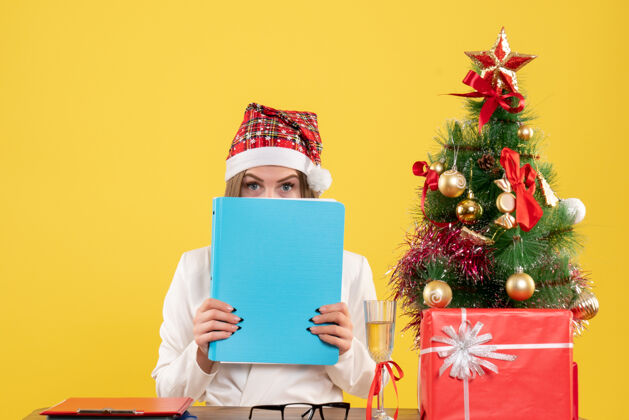 圣诞节前视图女医生坐在圣诞礼物举行的黄色背景上的文件庆祝医生坐着