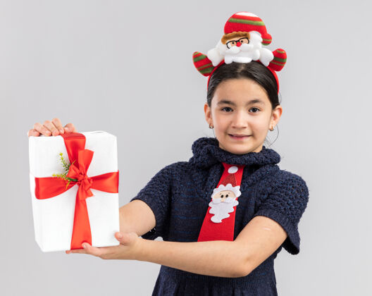 圈快乐快乐的小女孩 穿着针织连衣裙 头上系着一条红色领带 头上戴着一个有趣的圣诞圈 手里拿着圣诞礼物 脸上挂着微笑编织礼物头