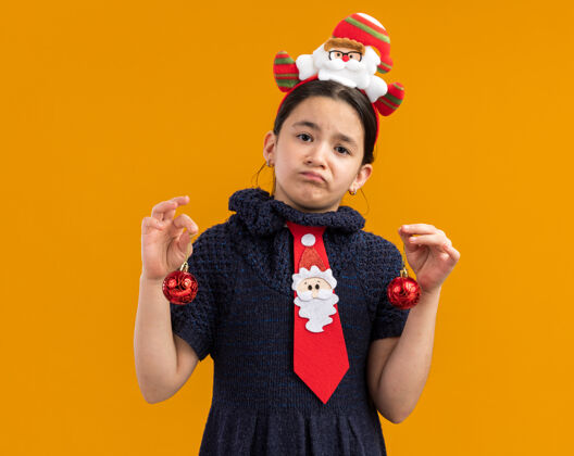 边缘穿着针织连衣裙的小女孩头上系着一条红领带 手里拿着圣诞球 神情困惑 表情悲伤编织小抱