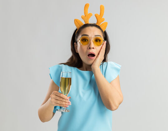 鹿身穿蓝色上衣的年轻女子戴着滑稽的鹿角边 戴着黄色眼镜 手里拿着一杯香槟 看上去既惊讶又惊讶新年眼镜杯子