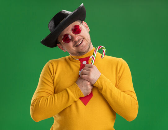 高领毛衣穿着黄色高领毛衣 戴着眼镜 打着滑稽的红领带 戴着黑色帽子 手里拿着糖果棒 脸上带着幸福的笑容站在绿色的墙上年轻人圣诞节手杖