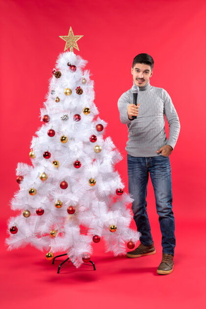 礼物新年气氛积极的家伙穿着牛仔裤站在装饰圣诞树附近圣诞节麦克风牛仔裤