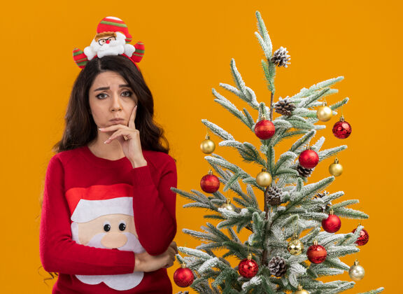 站着可疑的年轻漂亮女孩戴着圣诞老人的头带和毛衣站在装饰过的圣诞树旁 看着相机 手放在下巴上 橙色背景下的隔离树女孩圣诞老人