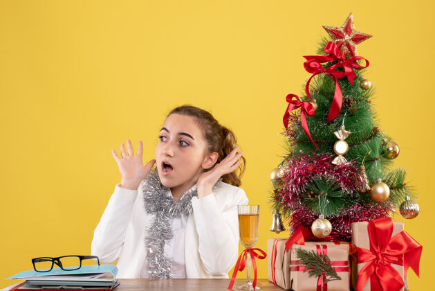 安排正面图：女医生坐在桌子后面 脸上带着恐惧 背景是黄色的圣诞树和礼盒花女医生脸