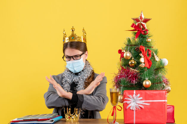 桌子正面图戴着面具戴着眼镜戴着皇冠的年轻女孩双手交叉着圣诞树和鸡尾酒礼物强化剂支撑穿着
