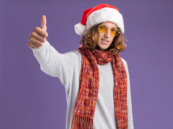 展示快乐的年轻人戴着圣诞老人帽 戴着黄色眼镜 脖子上围着暖和的围巾 脸上带着微笑 站在紫色背景上竖起大拇指 看着相机圣诞老人背景拇指
